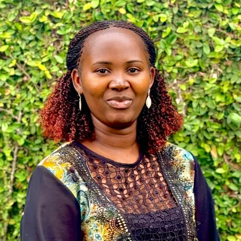 Milicent is wearing colorful, patterned top. She stands in front of a green leafy background, smiling subtly.