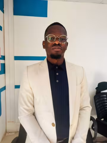 Muyunda is wearing a white blazer over a black shirt and glasses. He looks directly at the camera and stands against a white wall with blue stripes.