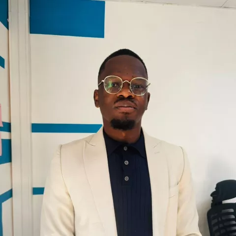 Muyunda is wearing a white blazer over a black shirt and glasses. He looks directly at the camera and stands against a white wall with blue stripes.