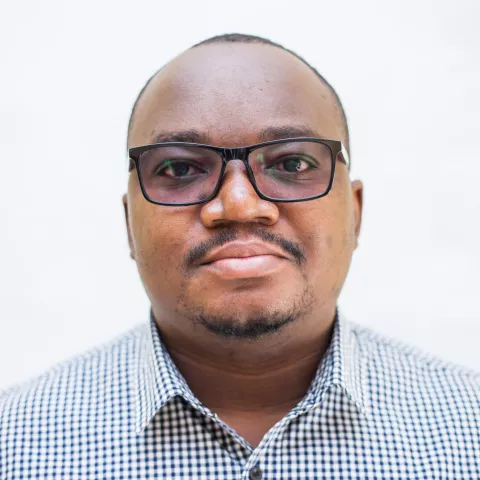 Mwanza is wearing a checkered shirt and glasses. The background is plain white.