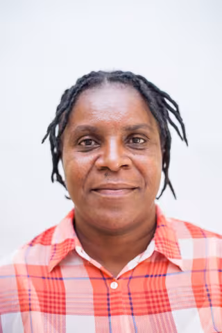 Nyambe is wearing a plaid red and white shirt. She is smiling. The background is plain white.