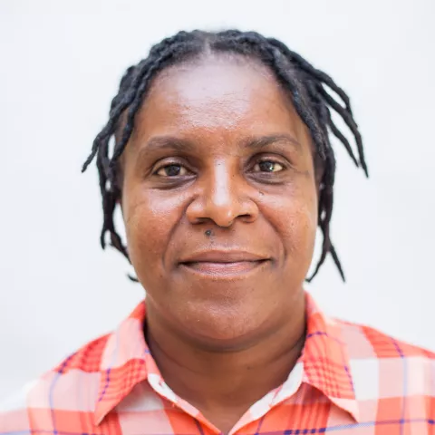 Nyambe is wearing a plaid red and white shirt. She is smiling. The background is plain white.