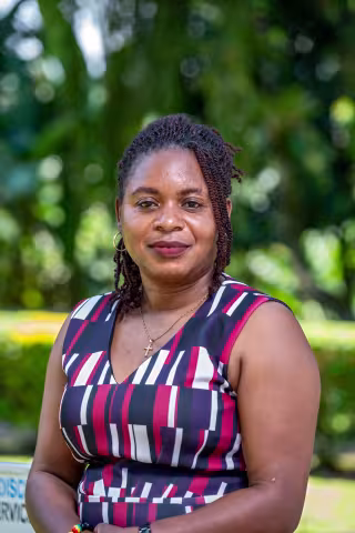 Paulah is wearing a patterned dress. She smiles slightly, standing against a background of blurred greenery.