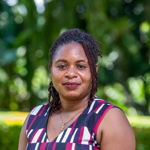 Paulah is wearing a patterned dress. She smiles slightly, standing against a background of blurred greenery.