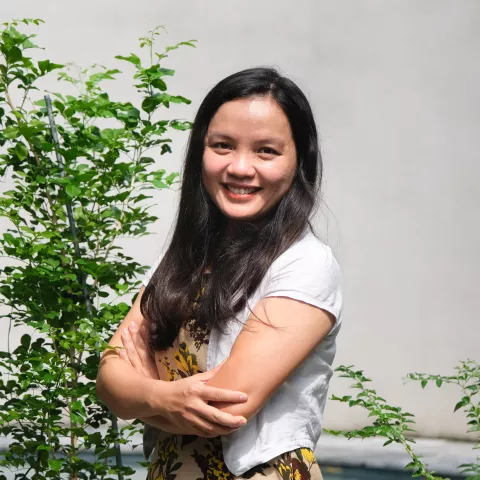 Phuong Ang is smiling with her arms crossed. She is wearing a white cardigan over a colorful patterned shirt. In the background, there are some green plants.
