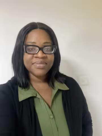 Precious is wearing glasses and a black cardigan over a green shirt. She is smiling against a white wall.