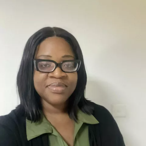 Precious is wearing glasses and a black cardigan over a green shirt. She is smiling against a white wall.