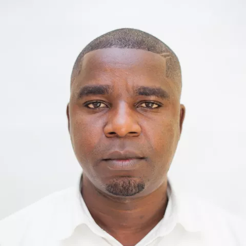 Prosper is wearing a white shirt as he looks directly into the camera. The background is plain white.
