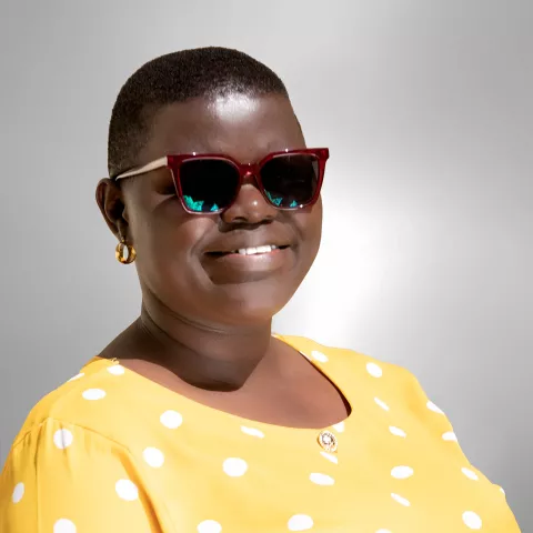  Rebecca is wearing dark red sunglasses and a bright yellow polka dot top. The background is a soft gray.