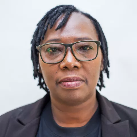 Sandra has a neutral expression. She wears glasses, a black blazer and black shirt. The background is plain white. 