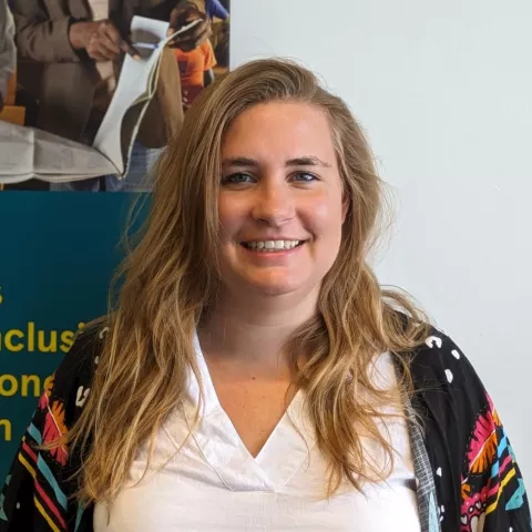 Sarah is wearing a white shirt and colorful patterned shawl. Background shows a partial poster with text. 