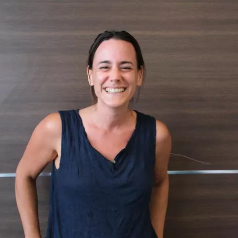 Silvija is wearing a blue top. She is smiling and standing in front of a brown wall. 