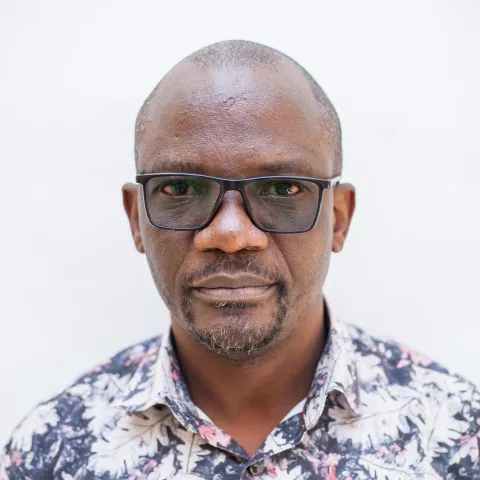 Siza is wearing glasses and a floral shirt. He stands against a plain white background, looking directly at the camera with a neutral expression.