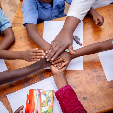 Six children's hands come together over a desk to demonstrate teamwork