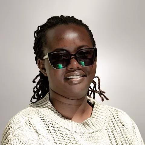 Vane is wearing sunglasses and a knitted sweater. She stands against a plain background. 