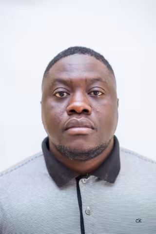 Vwambanji is wearing a polo shirt and looks directly into the camera. The background is plain white.