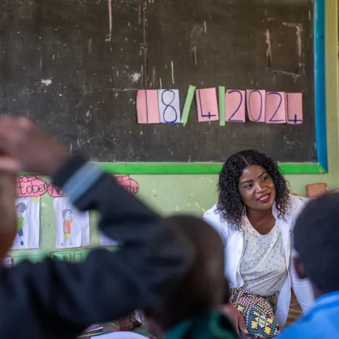 Teacher Emelda sits on the floor at the front of a classroom, speaking to a group of young children facing her.