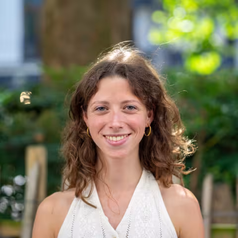 A smiling woman with shoulder-length curly brown hair stands outdoors wearing a sleeveless white top and gold hoop earrings. The background is softly blurred with green foliage, a tree trunk, and a wooden fence.