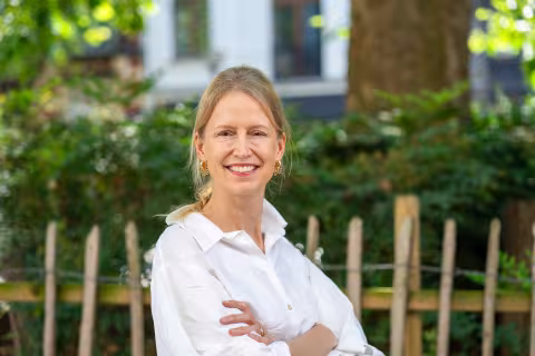 A smiling woman with blonde hair tied back stands outdoors with her arms crossed, wearing a white blouse, with a wooden fence and leafy trees softly blurred in the background.